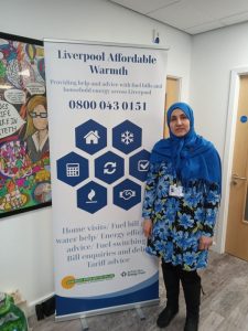 Our Community Energy Advisor, Djamila Serir, stands next to a banner displaying the name of our project, "Liverpool Affordable Warmth" and the telephone number to call our Save Energy Advice Line for support: 0800 043 0151.